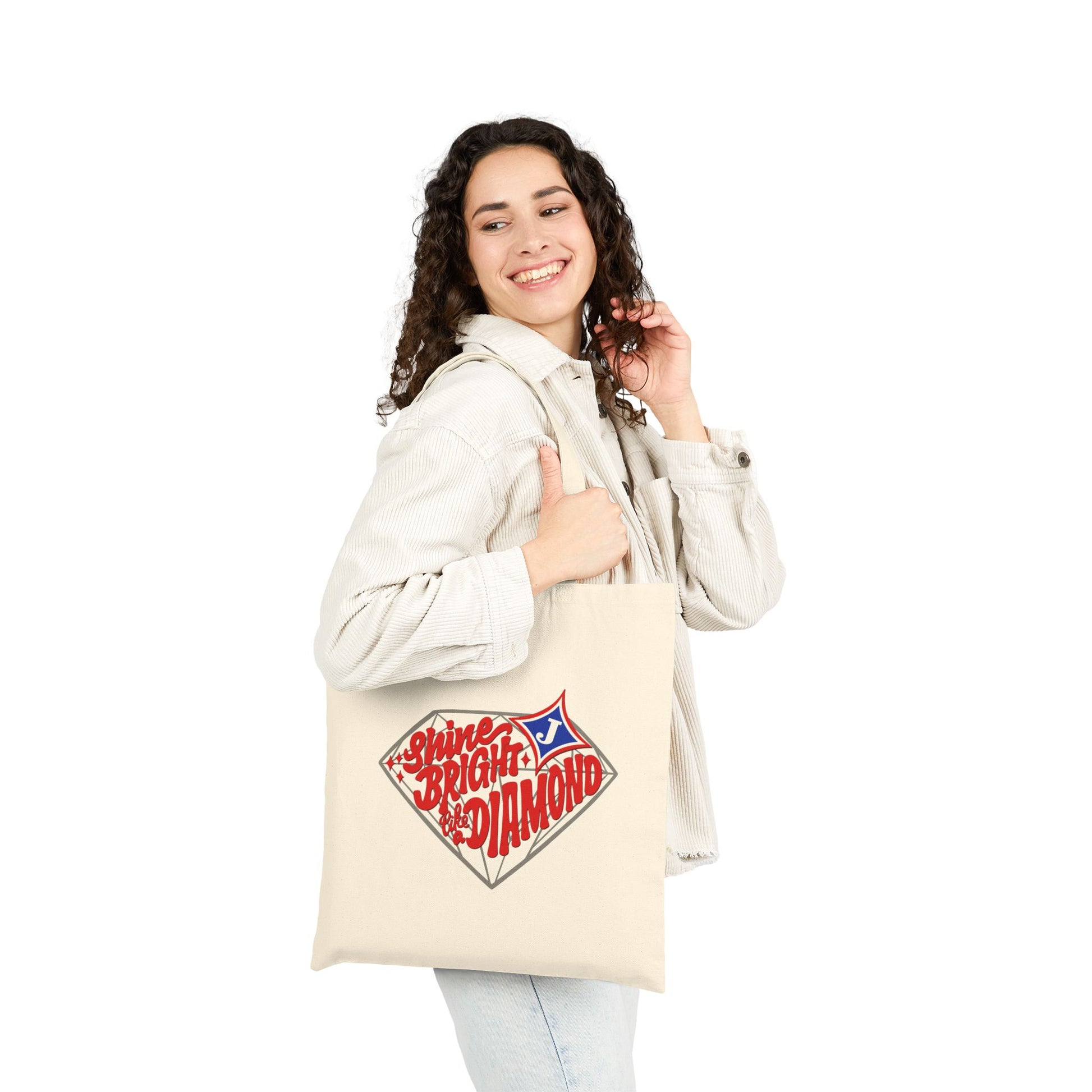 A woman wearing a light beige jacket and jeans holds a cream canvas tote bag with a red, white, and blue graphic that reads 'Shine Bright Like A Diamond’ with the Jefferson City schools logo